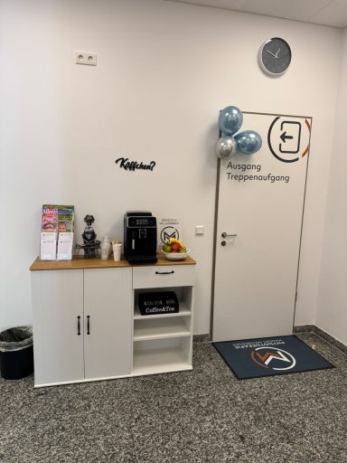Ecke mit Kaffeemaschine, Blumen und einer Tür, die zu einem Raum führt.